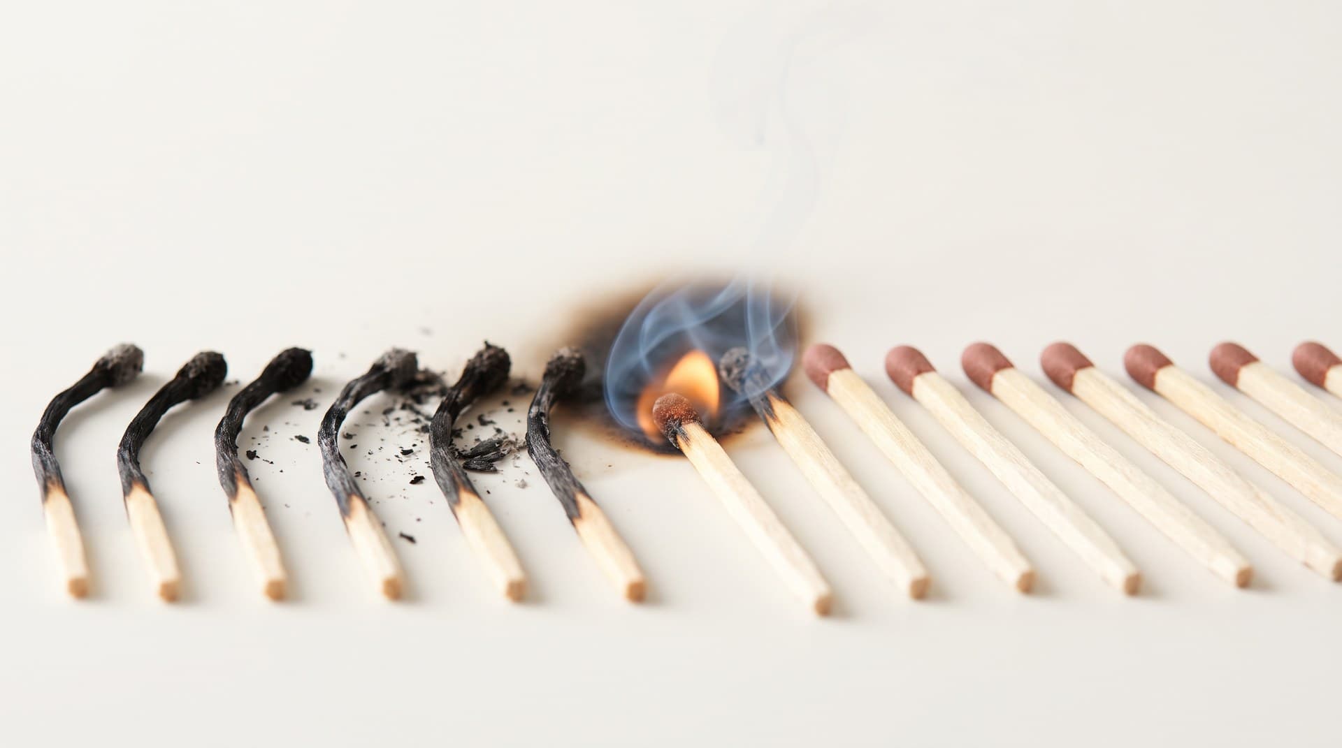 A row of identical wooden matches on a bone-white studio surface, six already burnt to black stubs, one mid-flame, the rest unused — a metaphor for every Claude Code GitHub Actions run starting cold and burning a fresh set of tokens