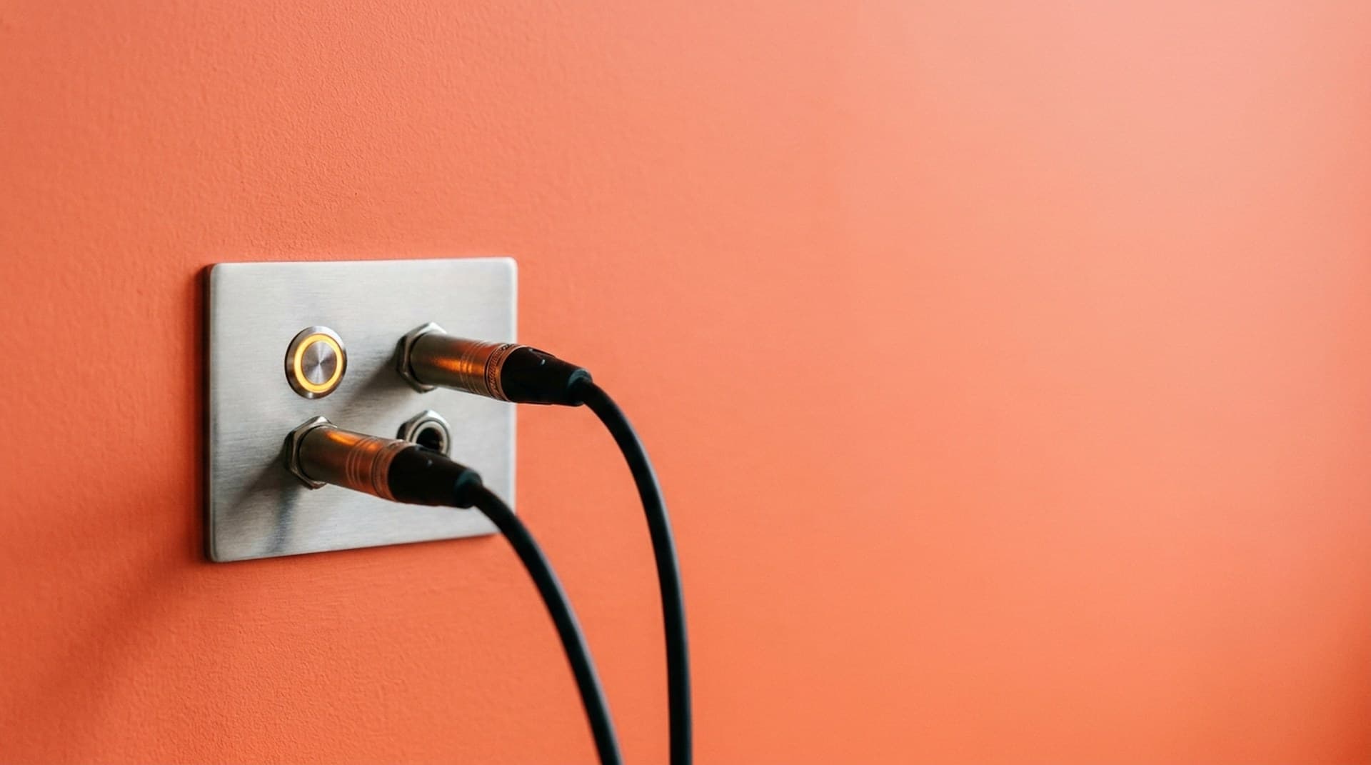 A brushed steel wall panel on a coral-orange wall with two black audio cables plugged in, one empty socket and one with a glowing amber ring, a metaphor for moving Eleven v4 Turbo to a different WebSocket endpoint