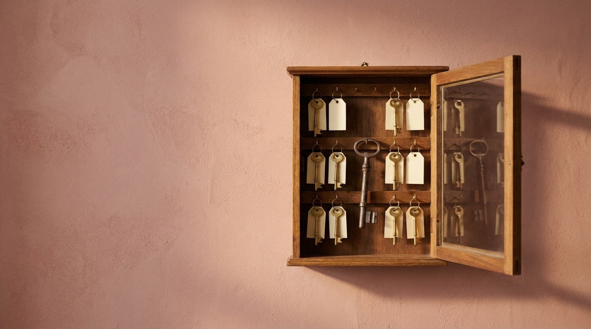 A vintage wooden key cabinet with its glass door half open on a dusty-rose plaster wall, rows of brass keys with blank tags around one oversized iron master key, a metaphor for n8n's $env access block and the N8N_ENCRYPTION_KEY it protects